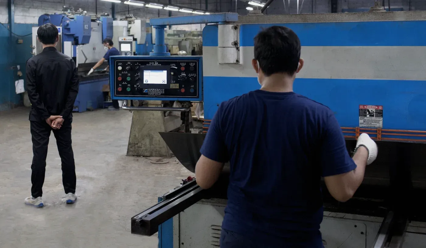 An operator at a CNC press brake on the Metosu factory floor while a supervisor watches